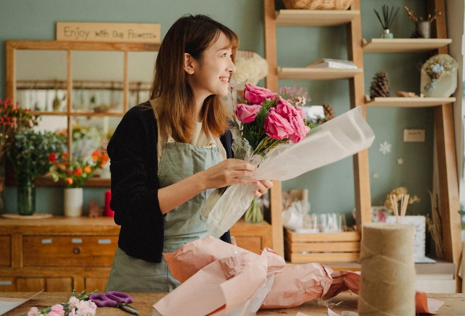 A smiling woman with brown hair, dressed in a light-colored apron over a dark cardigan, is holding a fresh floral bouquet wrapped in white tissue paper. The bouquet features vibrant pink roses and sma