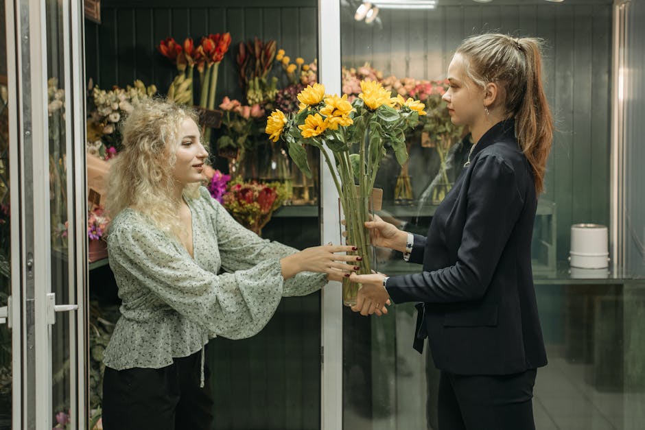 A florist at groveparkflowers.co.uk, dressed in a dark blazer, is handing a tall bouquet of vibrant yellow sunflowers and greenery to a customer with curly blonde hair, wearing a light green blouse, i