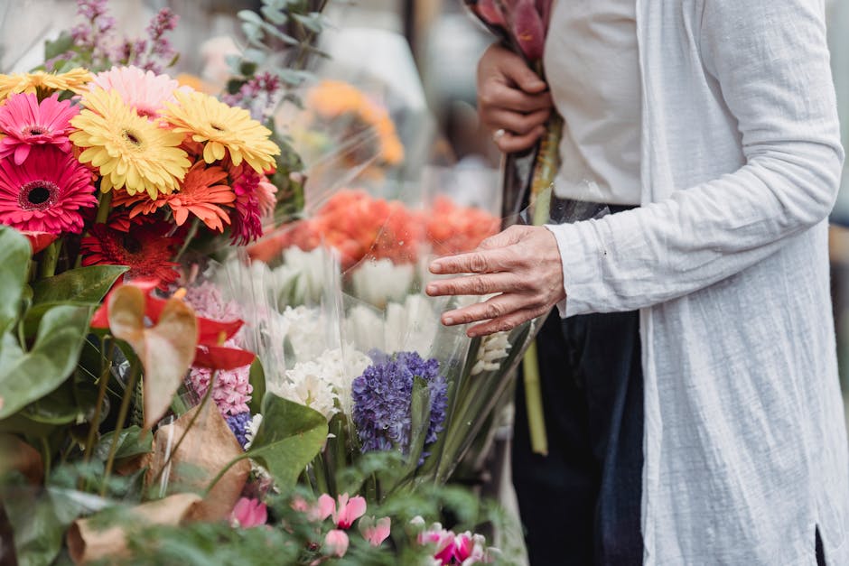 Close-up of a person selecting fresh flower bouquets at a market stall. The arrangement includes vibrant pink, yellow, orange, and purple flowers, such as daisies and hyacinths, wrapped in clear cello