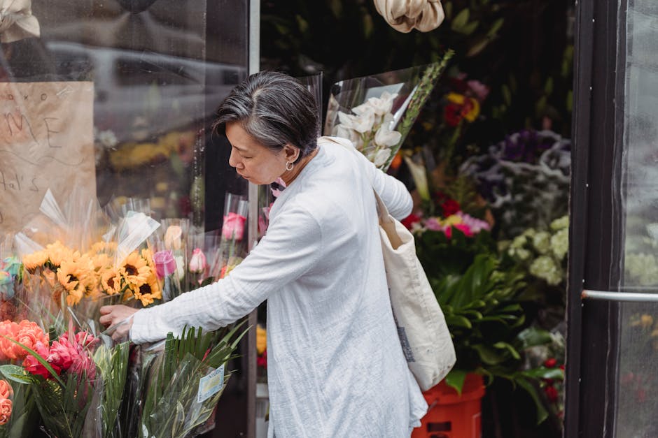 A mature woman with short, dark hair and wearing a white cardigan is selecting fresh flowers at an outdoor flower stall. She is reaching towards bouquets wrapped in clear plastic, featuring vibrant pi