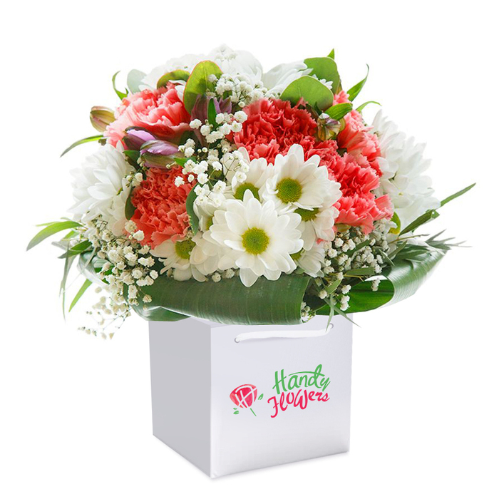 A lush, professionally arranged bouquet takes pride of place within a pristine white gift bag, its clean square silhouette setting off the vivid beauty of the flowers inside. The composition features soft coral and peach carnations layered with deeper red blooms, each petal ruffled and full, their colours harmonising like a Grove Park sunset. Interspersed are radiant white daisies with bold lime-green eyes, their cheerful faces reminiscent of meadows by the Thames Path. Delicate Gypsophila, or baby's breath, adds a light, airy element, its fine clusters echoing the gentleness of early morning dew. Along the edge, subtle purple-tinted buds-perhaps alstroemeria yet to open-peek out, suggesting growth and renewal. Ample deep green leaves and broad, glossy foliage wrap the bouquet, framing and supporting the blooms while ensuring freshness. The rounded form and thoughtful placement of every blossom reflect an expert touch, promising both visual impact and lasting vitality. The white gift bag, boasting strong ribbon handles for easy carrying, signals a bouquet meant for giving-suited for celebrations, milestone anniversaries, or simply to uplift someone in Grove Park or neighbouring Chiswick. The overall presentation-against a pure white background-emphasises both elegance and approachability, making this arrangement an inviting choice for those seeking same-day delivery of fresh, local flowers with a touch of Grove Park charm. Each detail speaks to the region's vibrant yet welcoming character.